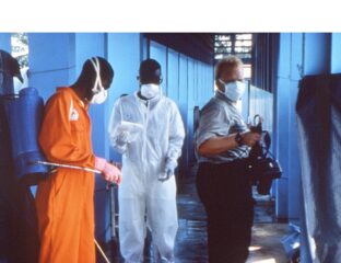 A man in an orange jumpsuit and two others in protective suits in an indoor setting.