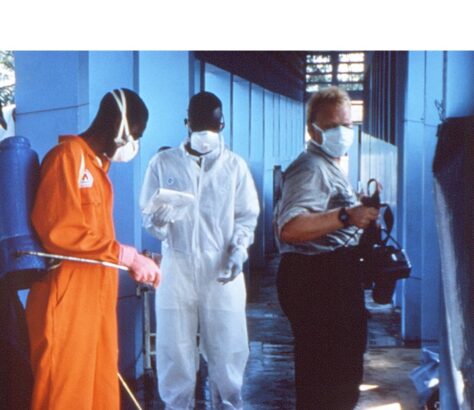 A man in an orange jumpsuit and two others in protective suits in an indoor setting.