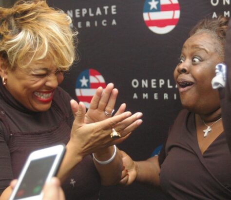 Two women joyfully high-fiving at an event with a OnePlate America backdrop.