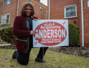 A woman poses with a campaign sign for Acqunetta Anderson in front of a brick house.