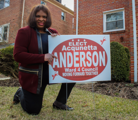 A woman poses with a campaign sign for Acqunetta Anderson in front of a brick house.