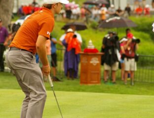 Golfer in orange shirt preparing to putt on the green with spectators in background.