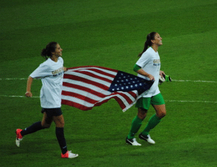 Two female soccer players running with an American flag on the field.