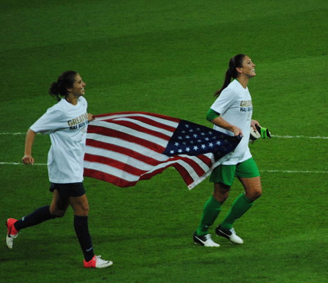 Two female soccer players running with an American flag on the field.