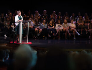 Woman speaking at a podium with an audience seated behind her on stage.