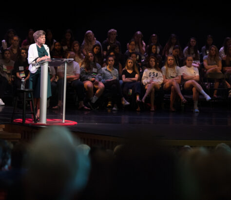 Woman speaking at a podium with an audience seated behind her on stage.