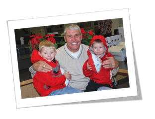 A man happily poses with two children dressed in red holiday outfits.