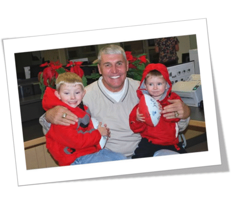A man happily poses with two children dressed in red holiday outfits.