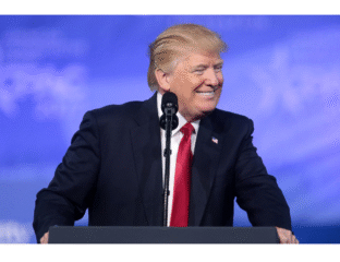 Donald Trump speaking at a podium with a blue sky background.