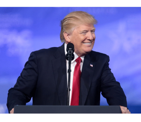 Donald Trump speaking at a podium with a blue sky background.