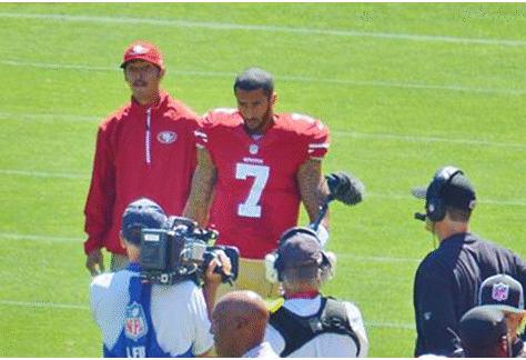 Two football players in red jerseys on the field surrounded by cameras and media.
