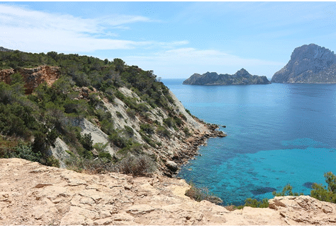 Rocky coastline with clear blue sea and distant islands under a bright sky.