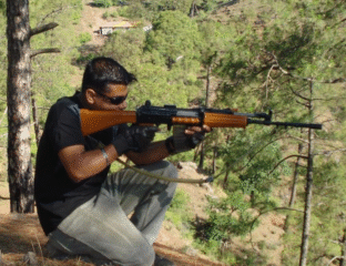 A man aiming a rifle outdoors in a forested area.