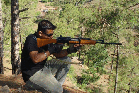 A man aiming a rifle outdoors in a forested area.