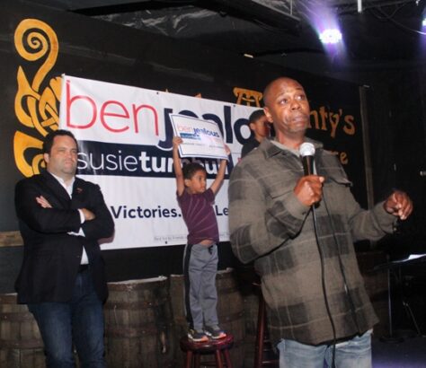 Man speaking at an event with a child and another man on stage.