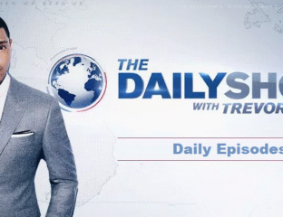 Host standing next to The Daily Show logo on a news set.