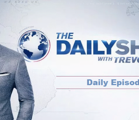 Host standing next to The Daily Show logo on a news set.