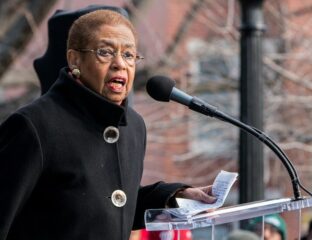 Elderly woman speaking passionately at a podium outdoors.