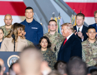 A group of people, including a man speaking, in front of a large American flag.