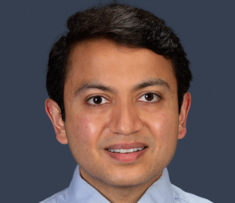 Professional headshot of a smiling man with dark hair in a light blue shirt.