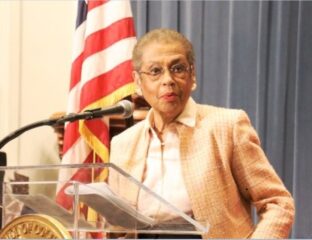 An elderly woman speaking at a podium with an American flag in the background.