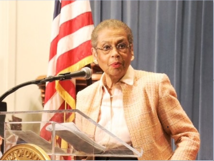 An elderly woman speaking at a podium with an American flag in the background.