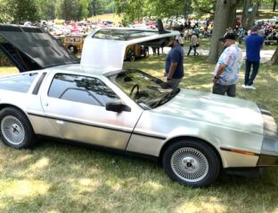 Silver DeLorean car with gull-wing doors open at a car show.