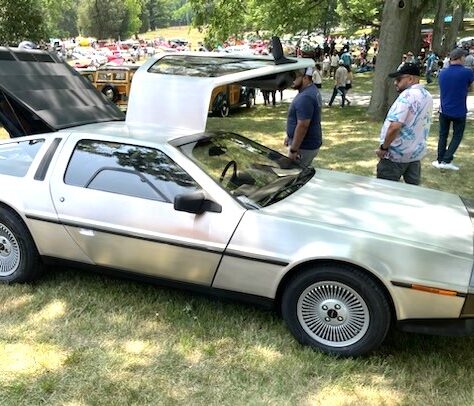 Silver DeLorean car with gull-wing doors open at a car show.