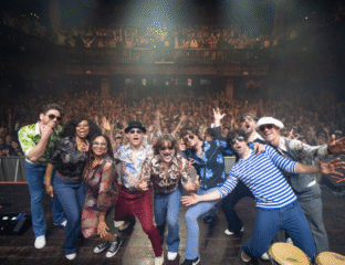 A lively group poses on stage with an enthusiastic crowd behind them.