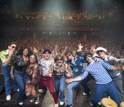 A lively group poses on stage with an enthusiastic crowd behind them.