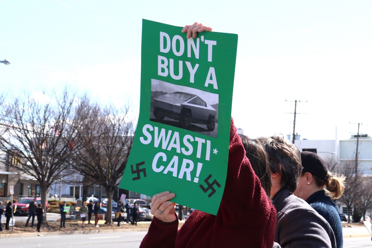 Protester holding a sign saying 'Don't buy a swasti-car!' with swastikas crossed out.