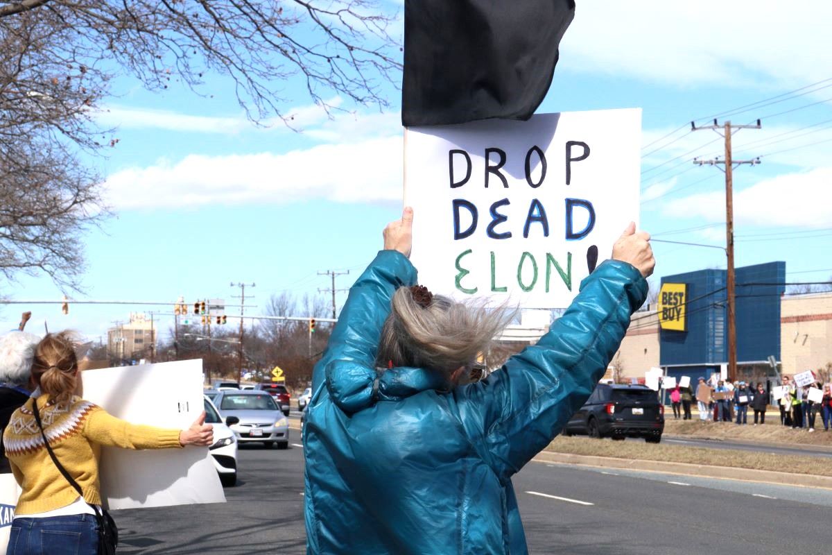 Person holding a sign that says 'DROP DEAD ELON' outdoors.