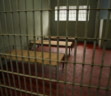 Empty prison cell with barred windows and basic wooden furniture.